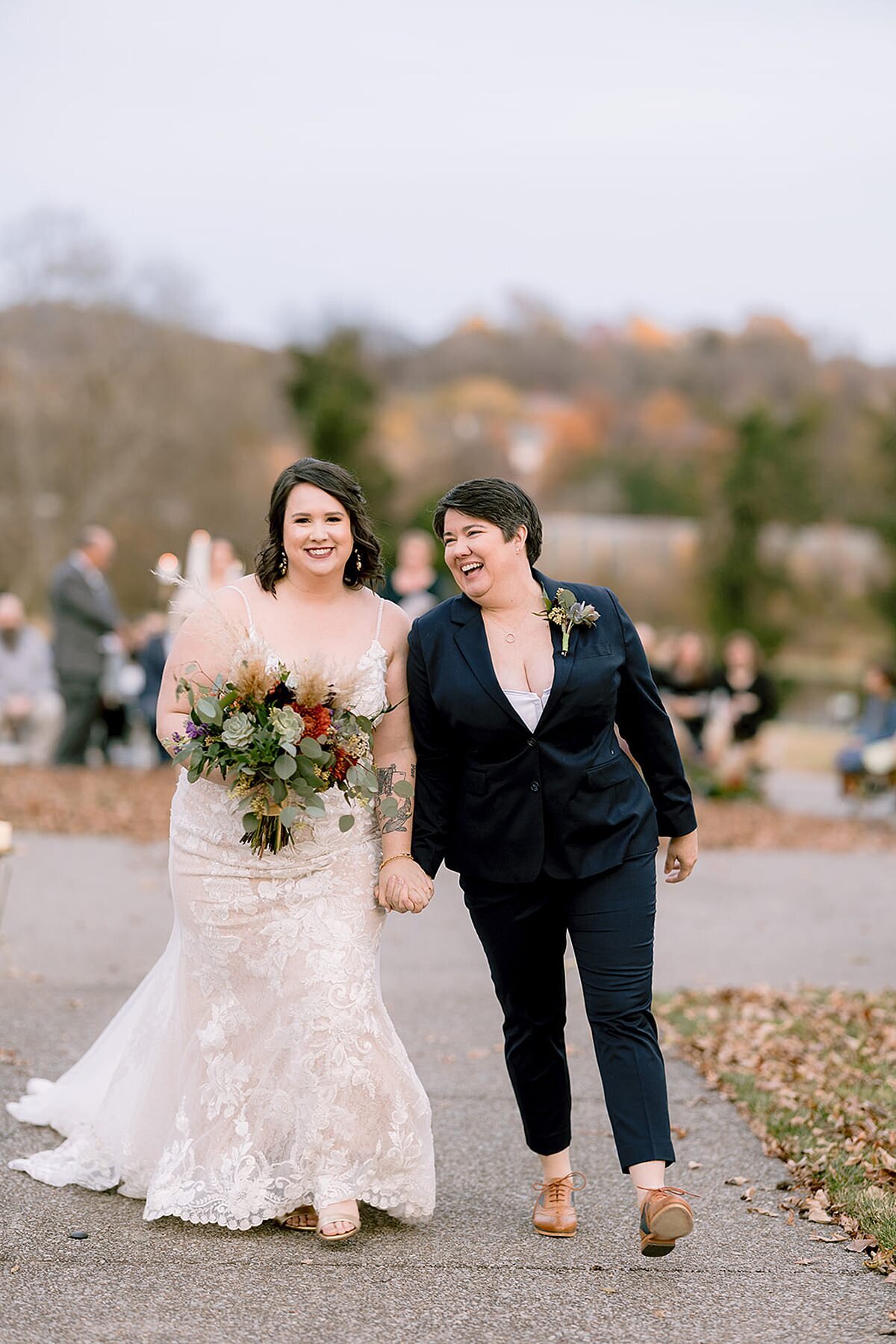 A bride wearing a white lace dress holding a bouquet of yellow, red and orange flowers walks hand in hand with her wife who is wearing a navy suit and small boutonniere of greenery