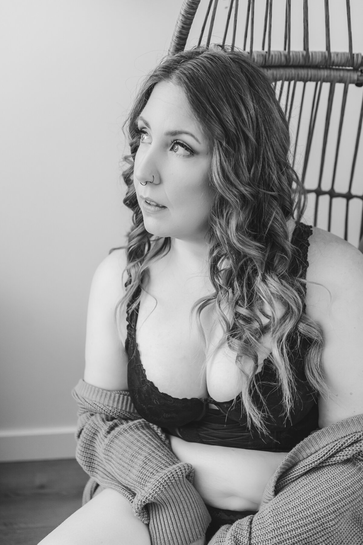 A woman wearing a black bra and a mustard cardigan, sitting in a wicker chair, gazing off in contemplation.