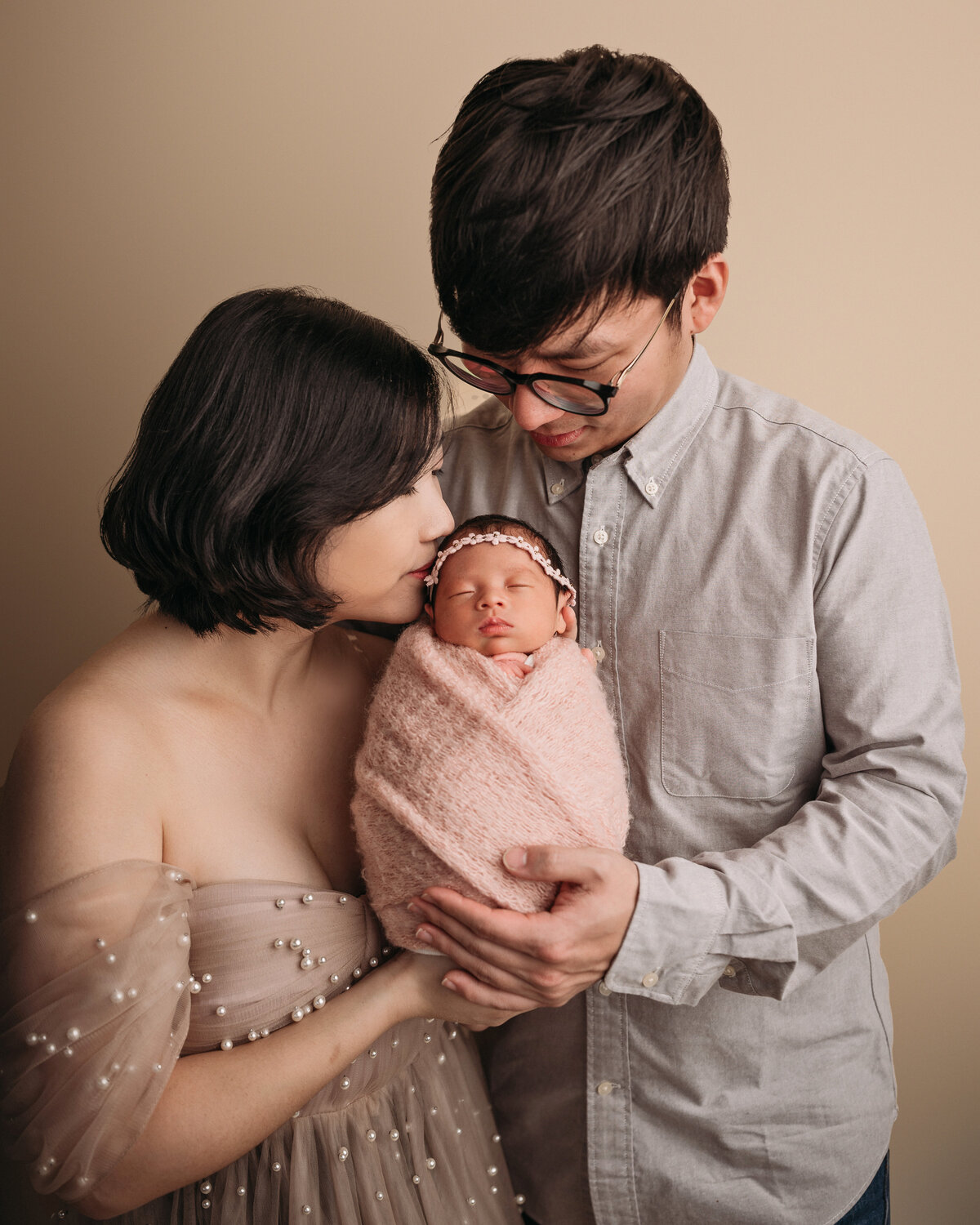 new mom wearing a cream pearl gown kissing newborn baby girl  standing next to dad wearing glasses and grey shirt while they both hold their brand new baby girl swaddled in pink and wearing floral headband in front of cream backdrop