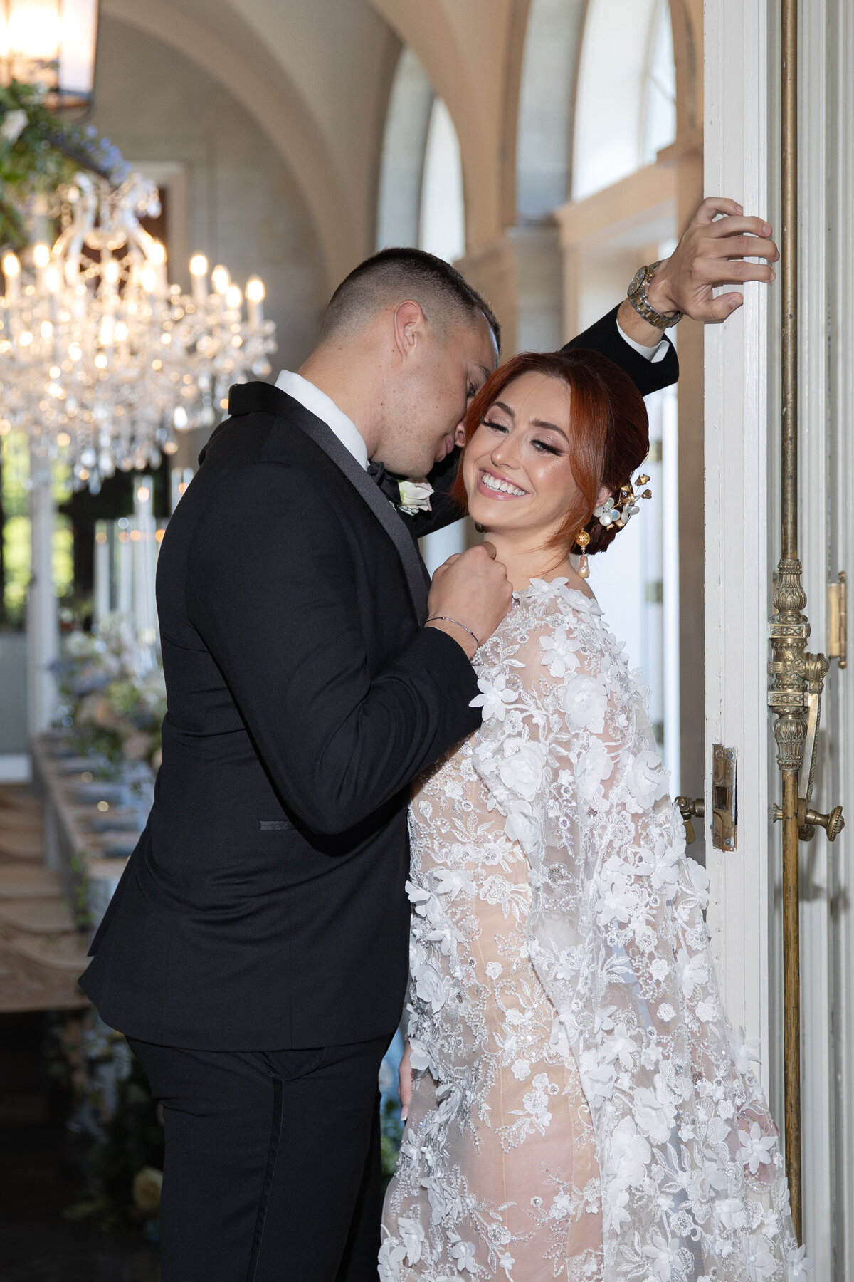 Elopement Wedding Packages Rhode Island | Kelsey Sheehan Photography Timeless Rhode Island Weddings | A couple lovingly embraces near an ornate door. The man in a black suit leans close, while the woman in a floral lace dress smiles, exuding joy. A chandelier softly illuminates the elegant setting.