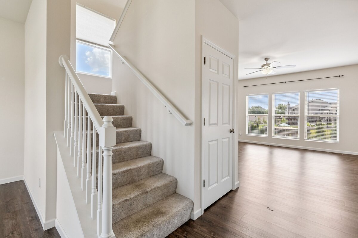 Real estate photo of a home showing the stairway, and leading into a living room with large windows looking out the back.