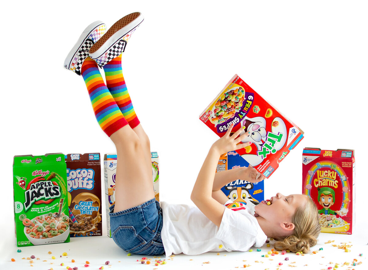 Little girl laying on white backdrop pouring cearel in her mouth wearing rainbow socks and rainbow vans.