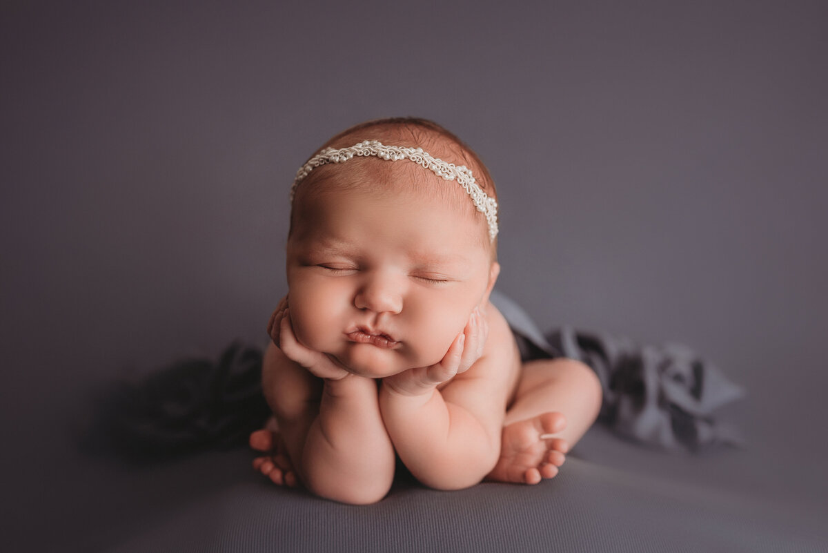 2 week old newborn girl asleep posed on elbows with chin on hands and white pearl headband on blue fabric backdrop