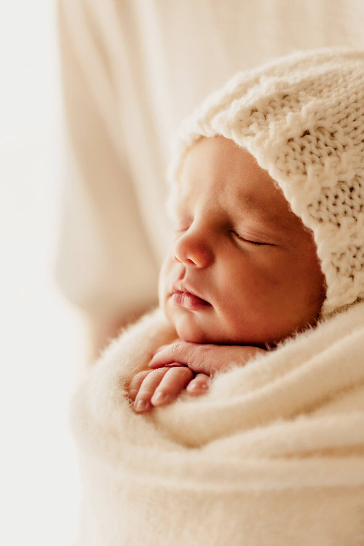 Newborn baby girl with her hands under her chin wearing a white knit bonnet for a profile photo.