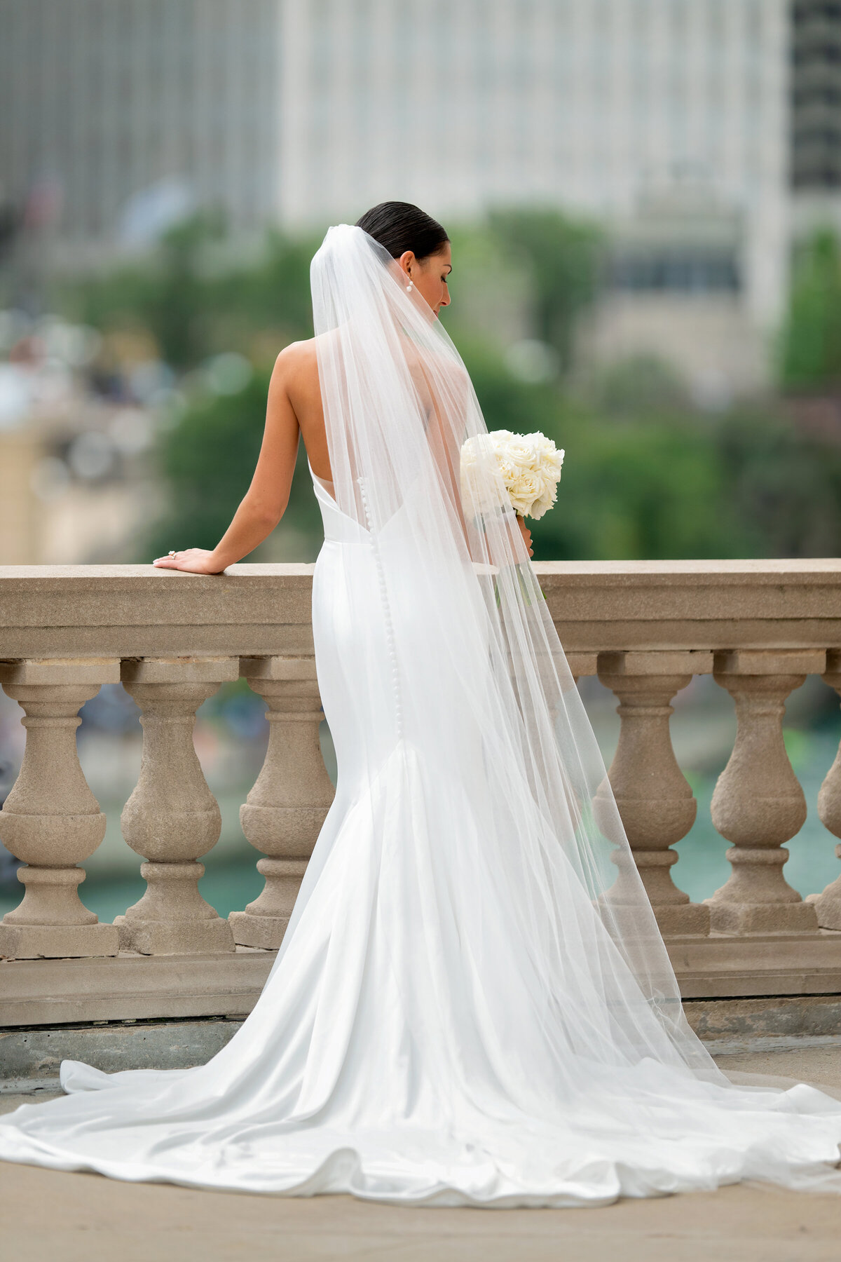 Bride in white wedding dress and long cathedral veil showing her back while leaning on pillar railing  next to Chicago River