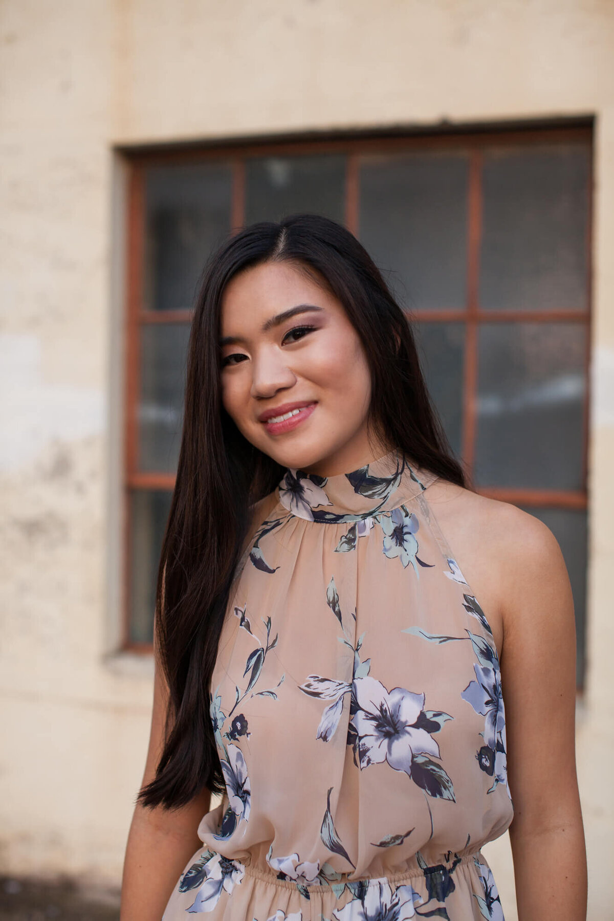 A lovely Asian American teen wearing a blush and blue floral jumper in front of industrial windows. Captured by Springfield, MO senior photographer Dynae Levingston.