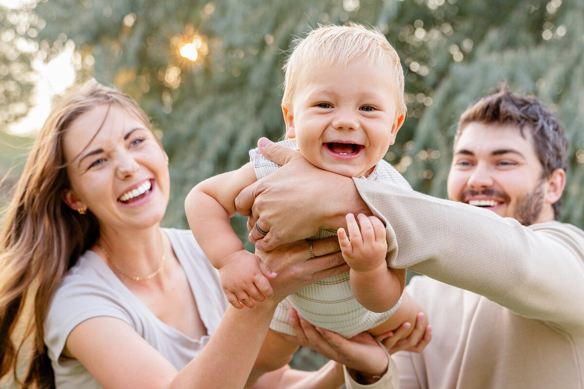 Close-up of a baby’s joyous expression as parents hold him up, captured in golden sunlight.