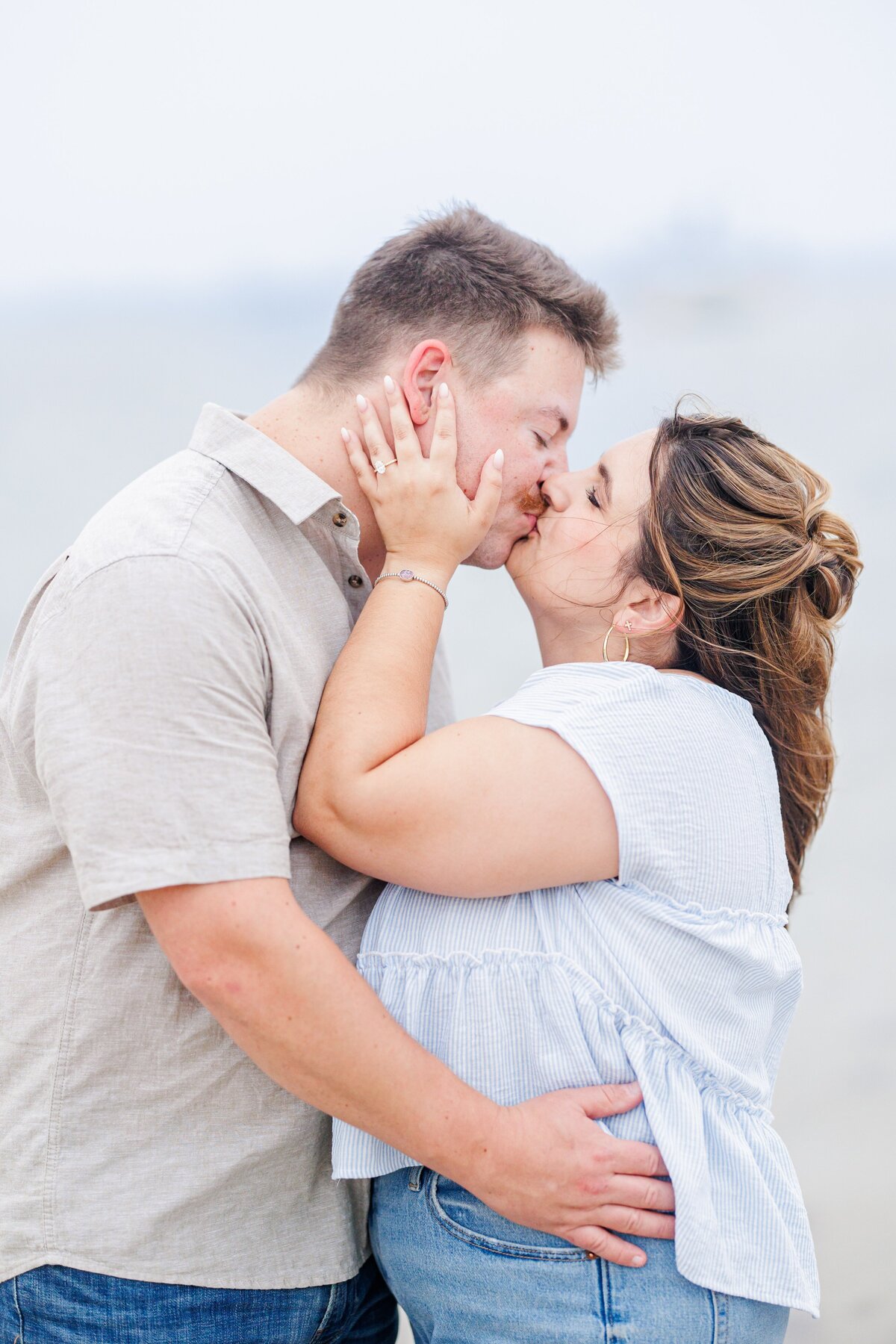 Close-up of a couple kissing on the beach, the woman’s hands on the man’s face showing her engagement ring, with a soft ocean backdrop at Chatham Bars Inn in Chatham, Massachusetts — representing the portfolio of a Cape Cod engagement photographer.