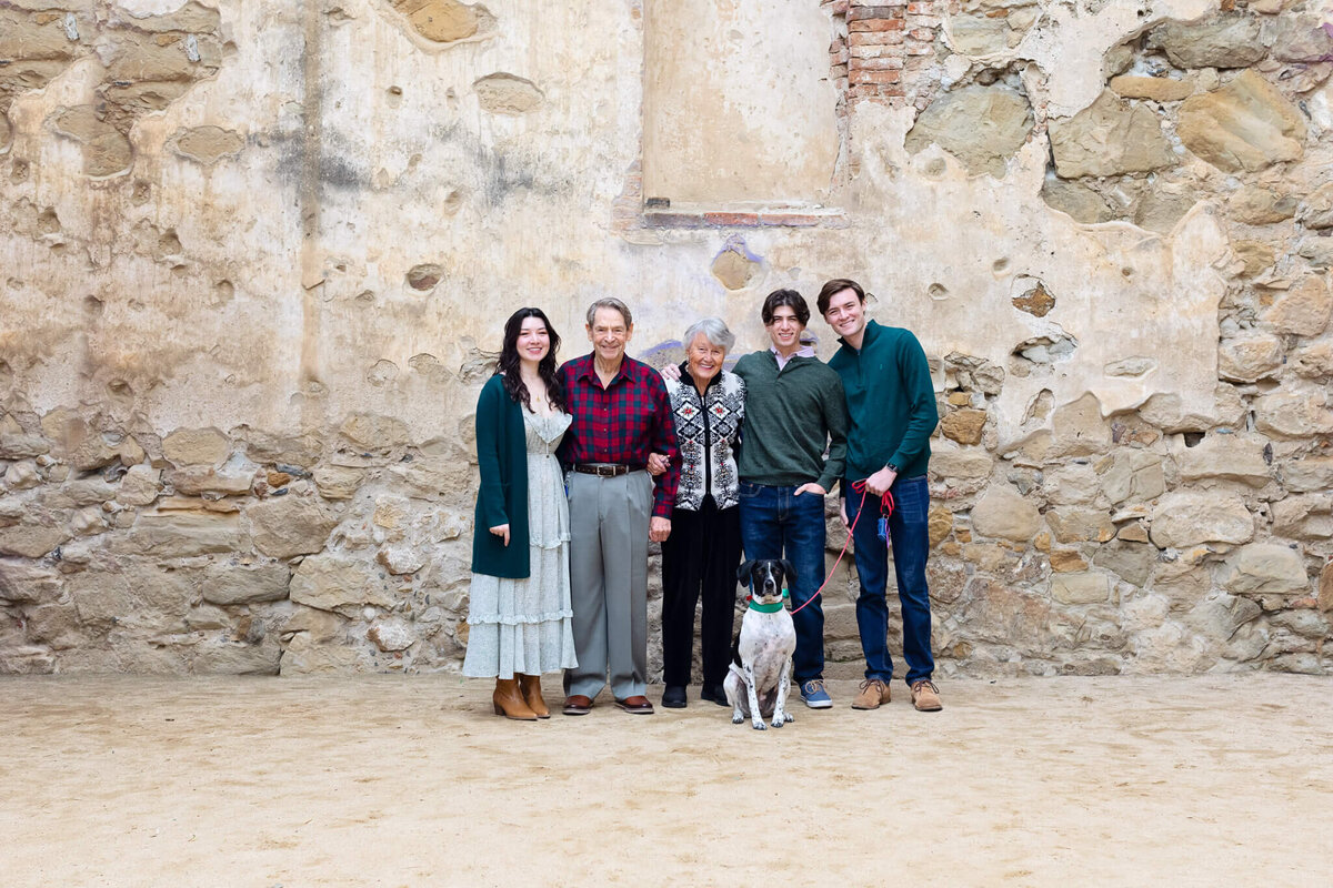 Three young adult kids and their grandparents and dog smiling in front of a rock wall at San Juan Capistrano.