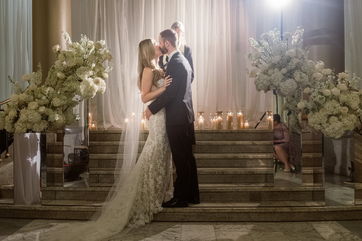 A bride and groom kiss surrounded by white flowers on the marble steps of Windsor Ballrooms.
