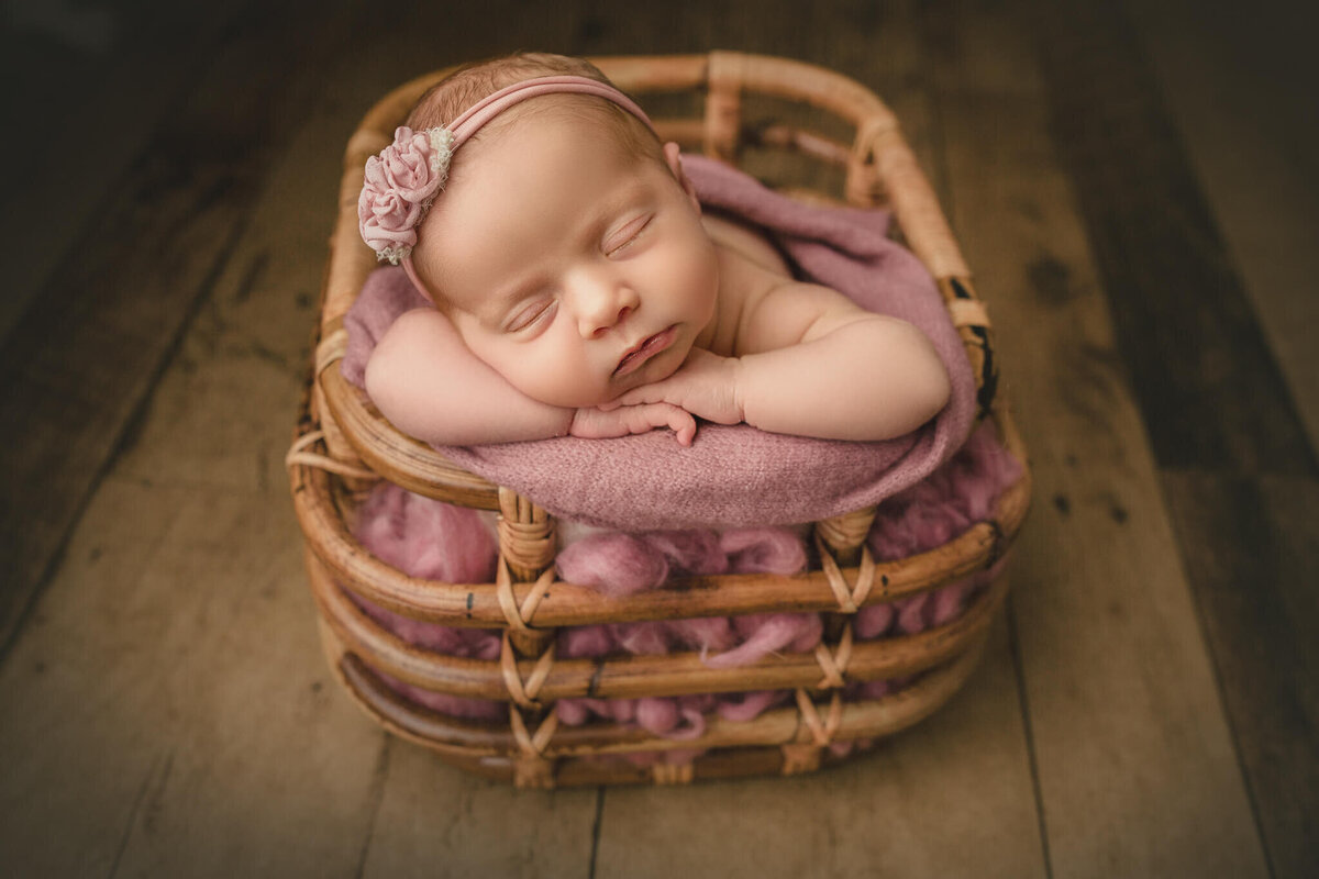 baby girl resting with chin on hands in bamboo basket on dark wood background in Hamilton, ON newborn photography studio