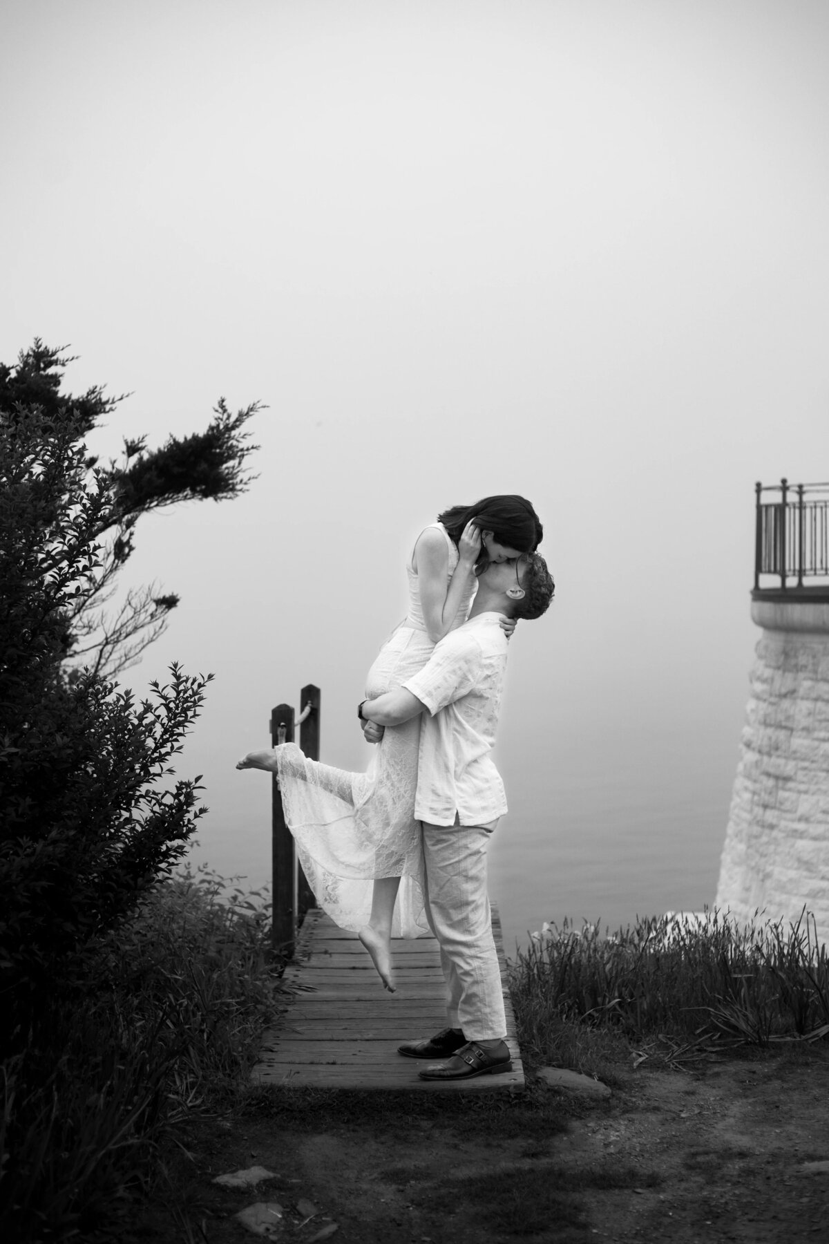 Wedding Photographers in RI | Kelsey Sheehan Photography Timeless Rhode Island Weddings | A couple shares a joyful kiss on a wooden walkway by a foggy waterfront. The man lifts the woman, surrounded by greenery and a stone railing. Black and white.