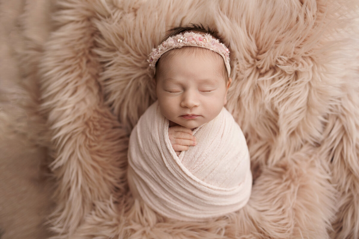 Newborn baby swaddled in a light pink blanket, sleeping peacefully on a fluffy pink rug, wearing a pink floral headband.