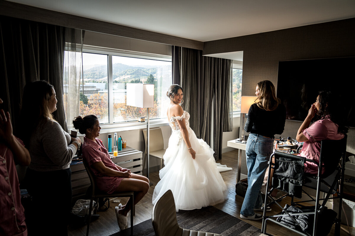 A heartfelt moment as the bride reveals her stunning wedding dress to her bridesmaids in the elegant setting of the AC Hotel Pleasanton. The bridesmaids' reactions are filled with excitement and joy, perfectly capturing the anticipation and love shared during this special getting-ready moment. A timeless memory from a beautiful Northern California wedding day.