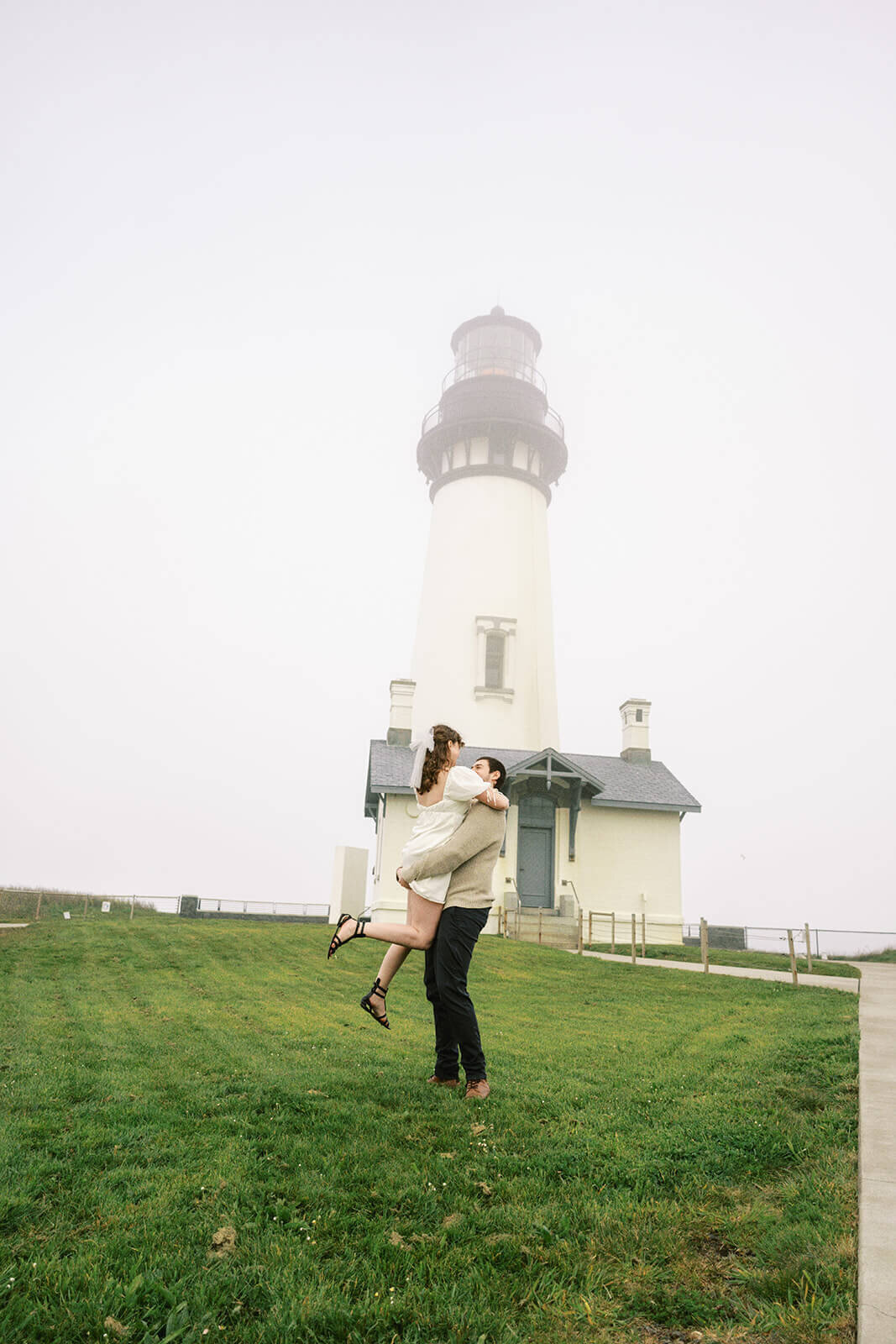 A couple embrace with Yaquina Head Lighthouse in the background. 