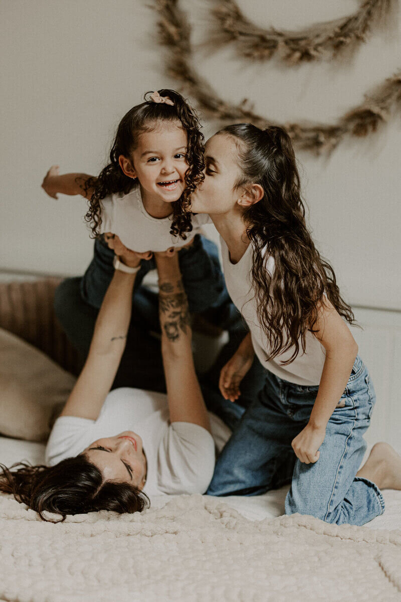 Maman faisant l'avion avec une de ses filles pendant que l'autre l'embrasse sur la joue dans un décor cosy à mon studio photo en Vendée.