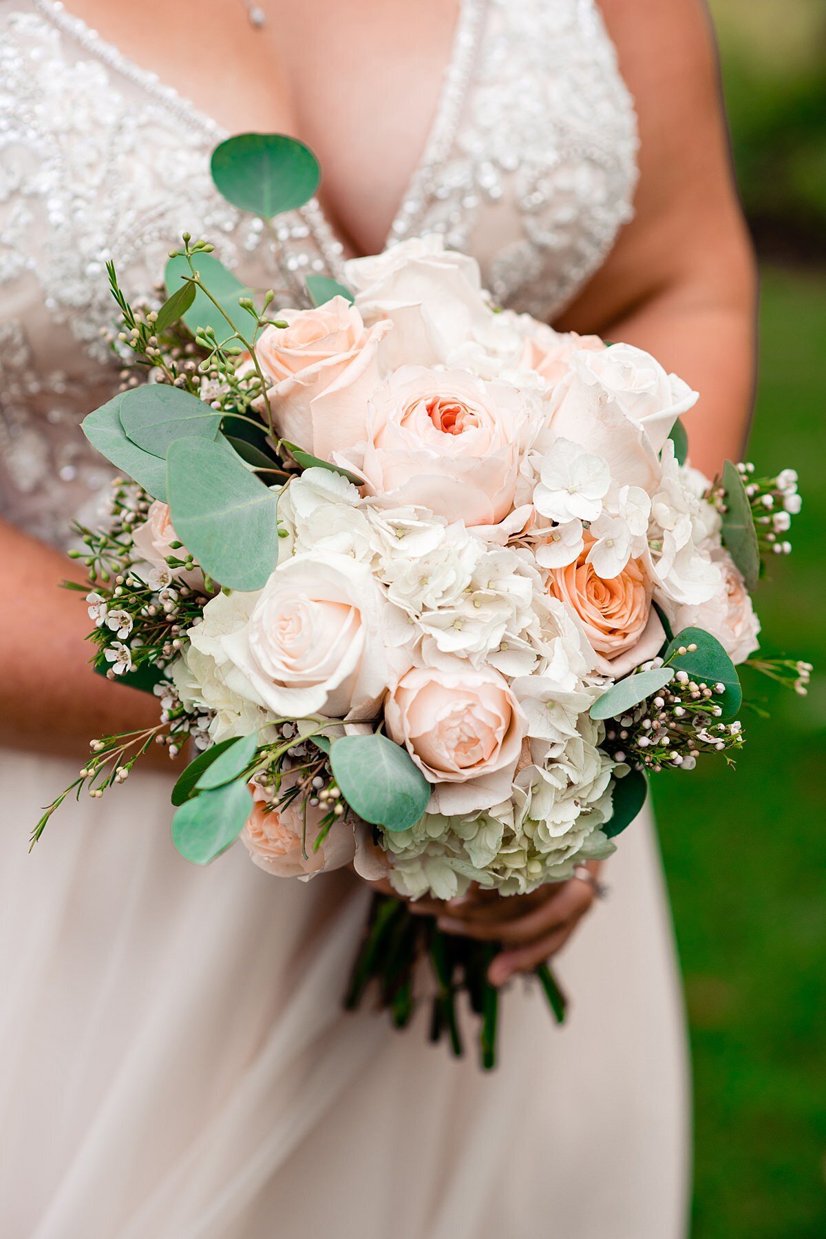 A bride wearing a wedding dress with a beaded bodice and deep v-neck holds a bouquet of white hydrangea, blush roses baby's breath and eucalyptus.