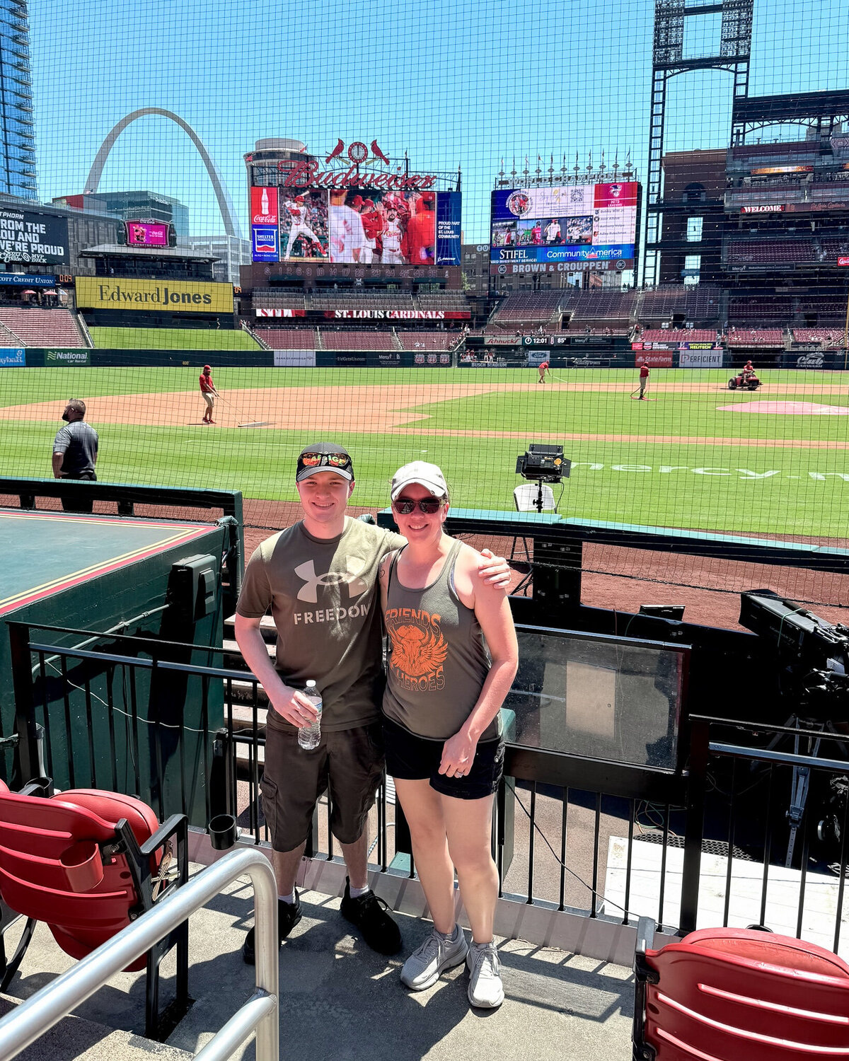mom and son standing in a baseball stadium with the players warming up behind them, smiling at the camera with arms around each other