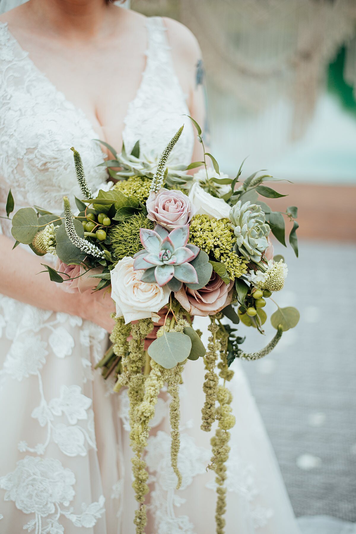 A cascading boho bridal bouquet with ivory roses, blush roses, green succulents, green carnations, silver dollar eucalyptus, white veronica and long cascades of green flowers. The bride is wearing an ivory lace wedding dress with a plunging neckline.