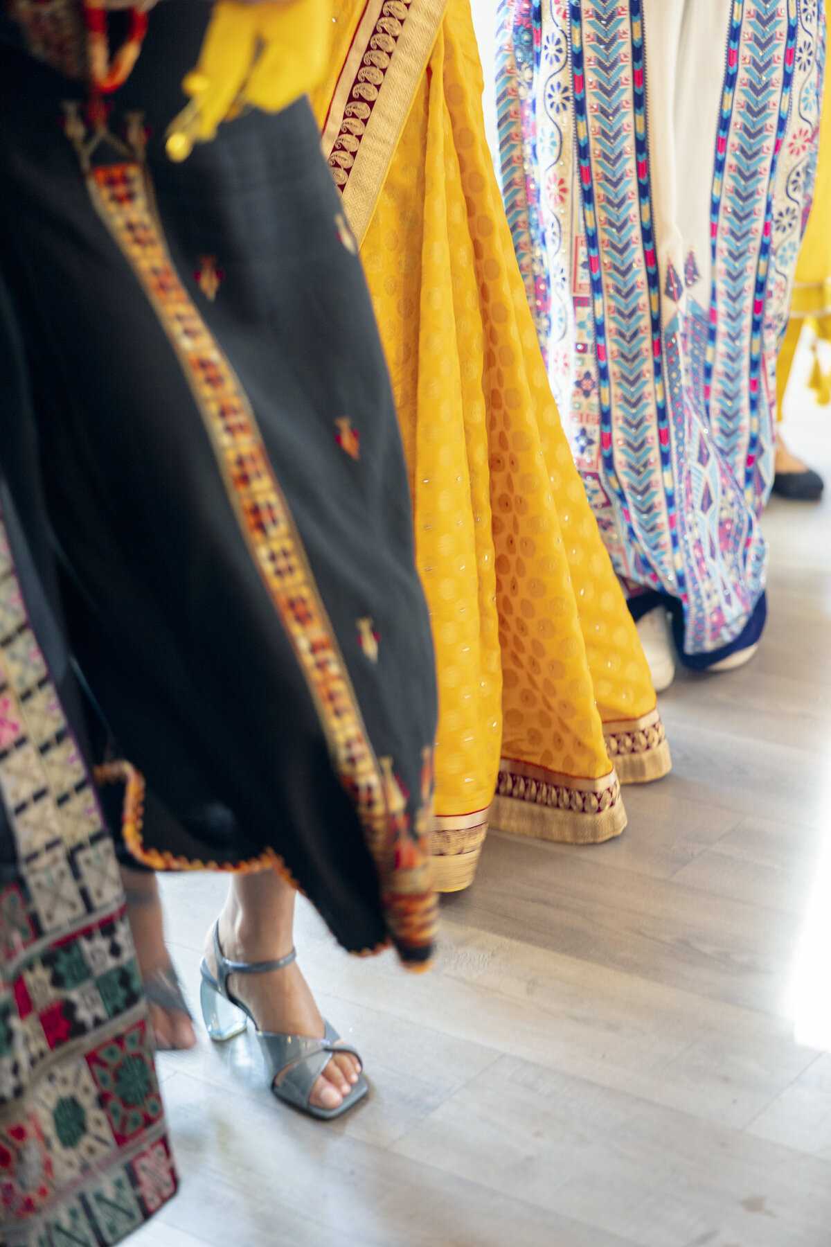 Women dressed in colorful traditional Indian sarees, including black, yellow, and multicolored patterns, walk indoors. The focus is on the vibrant fabric and flowing skirts, with one wearing silver sandals.