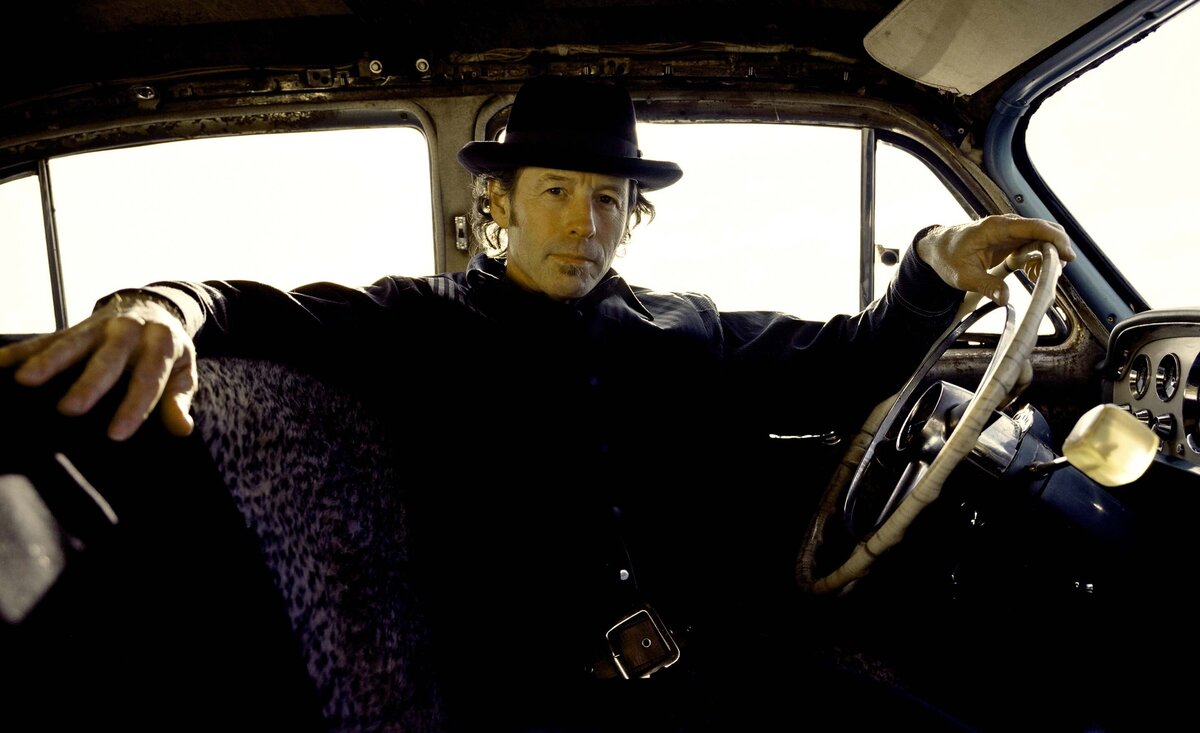 Male musician photo  Barney Bentall wearing black shirt black cowboy hat sitting against door inside old car