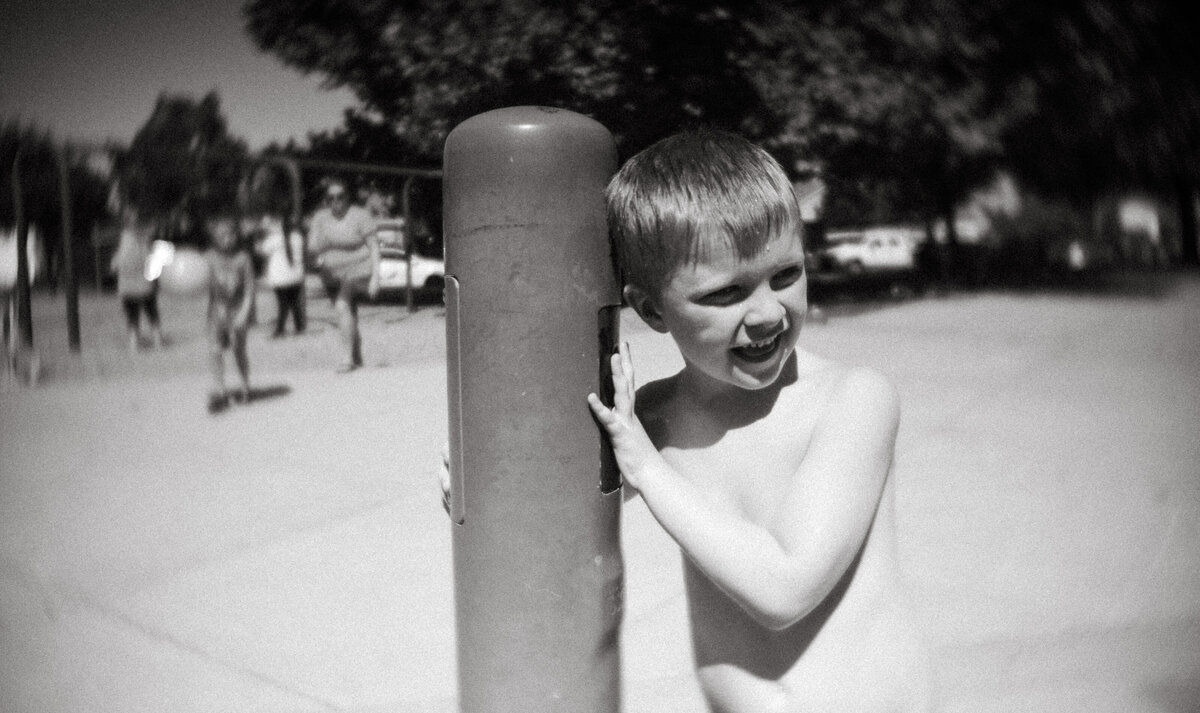 a little boy stands against a pole at a park, starting the water feature and ready for excitement