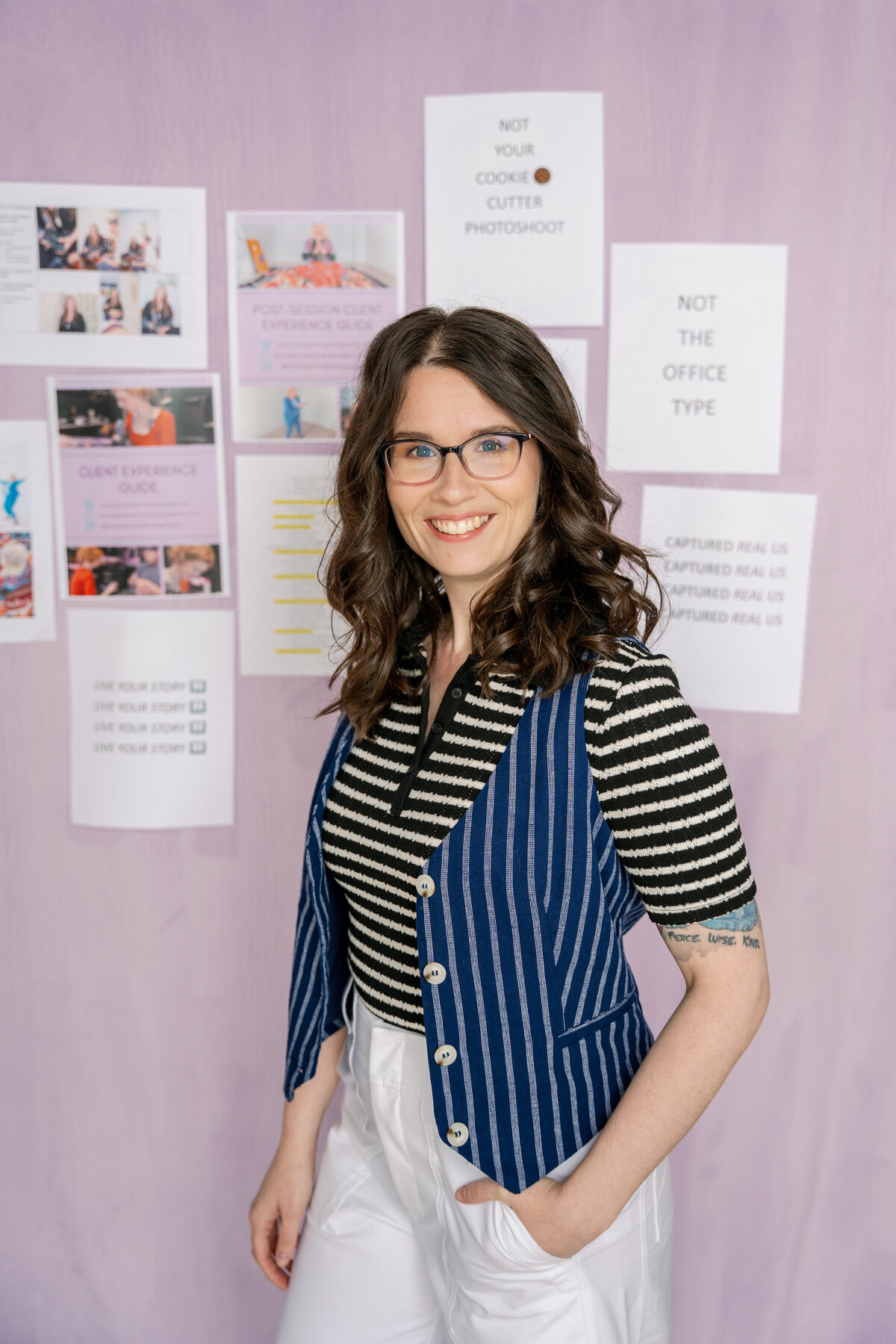 Alicia, lead Brand Photographer standing in front of a light purple background with printouts of her business assets pinned behind her. She is smiling at the camera with one hand in her pocket