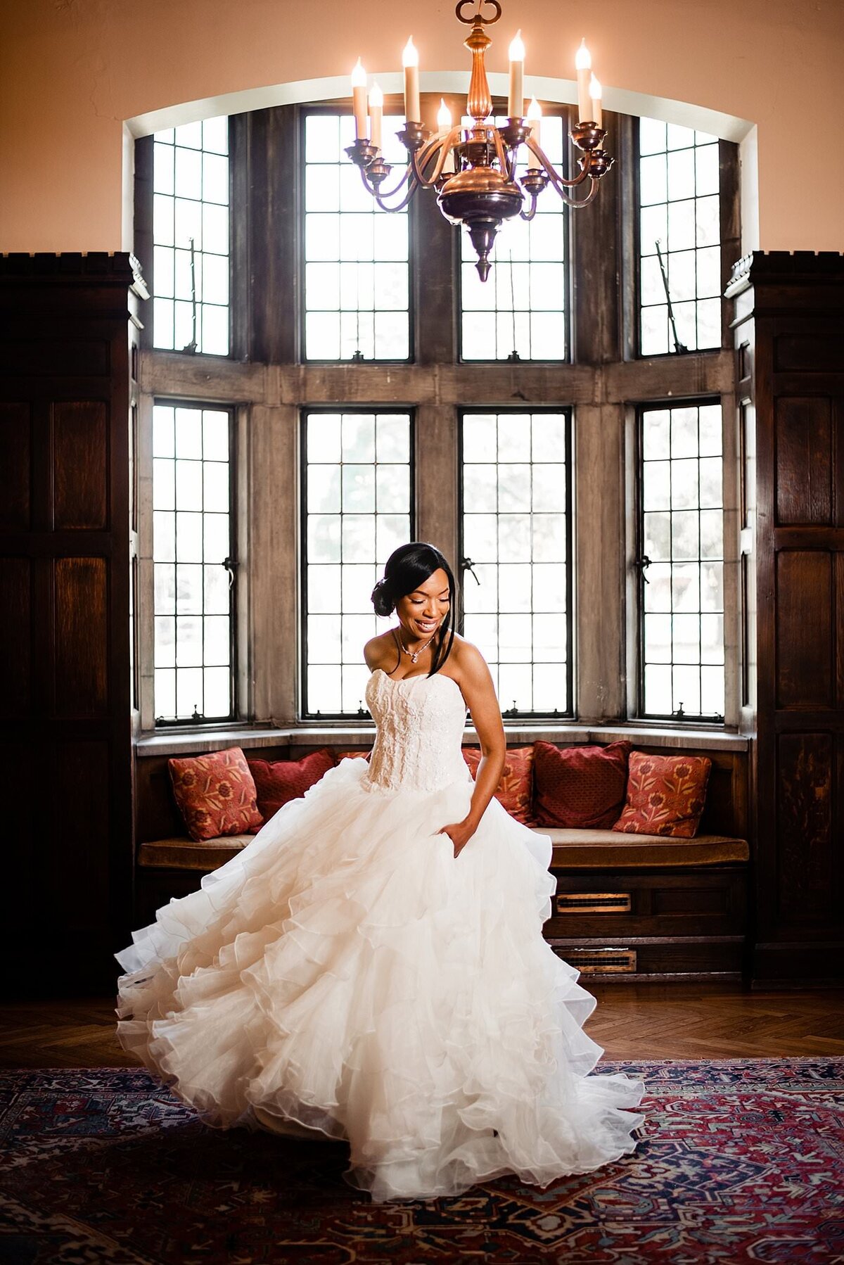 The bride, wearing a strapless dress with a fitted bodice and very full ruffled skirt twirls excitedly in front of a massive bay window.