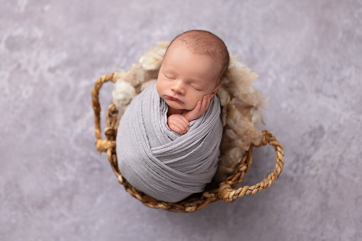 Newborn baby boy wrapped in soft grey fabric curled inside a woven basket on a neutral backdrop.