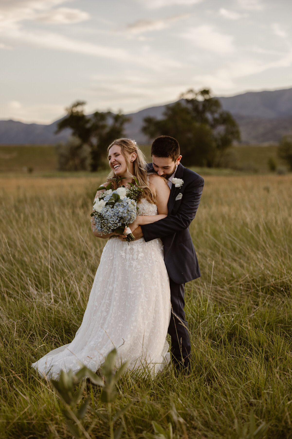 Bride laughs joyfully while holding a bouquet of hydrangeas as her groom embraces her from behind in a grassy field at sunset with mountains in the background.