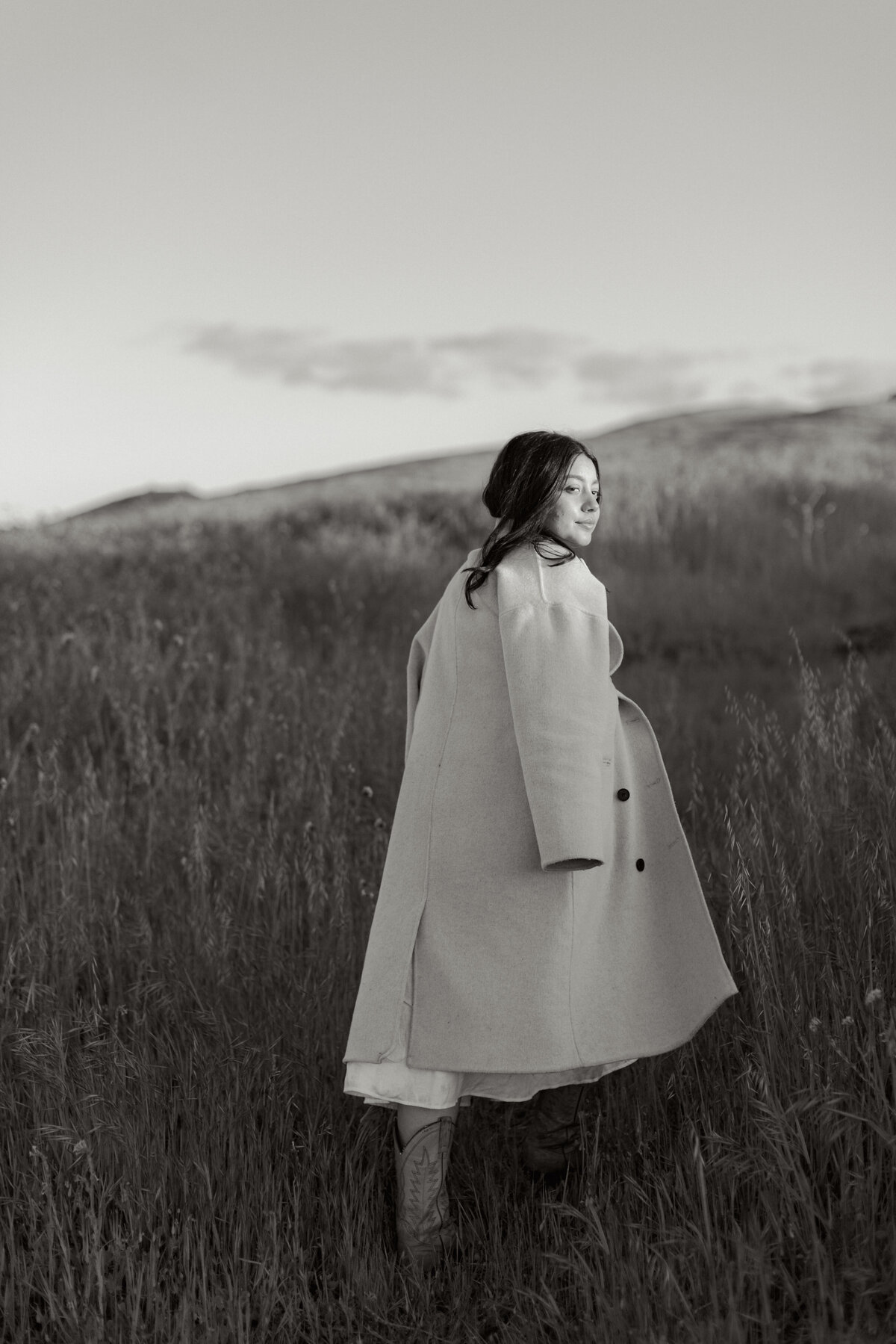 Black and white image of a woman walking through tall grass in Greenville, SC. She wears a long coat over a dress and cowgirl boots. Editorial-style portrait by Dani Lacey.