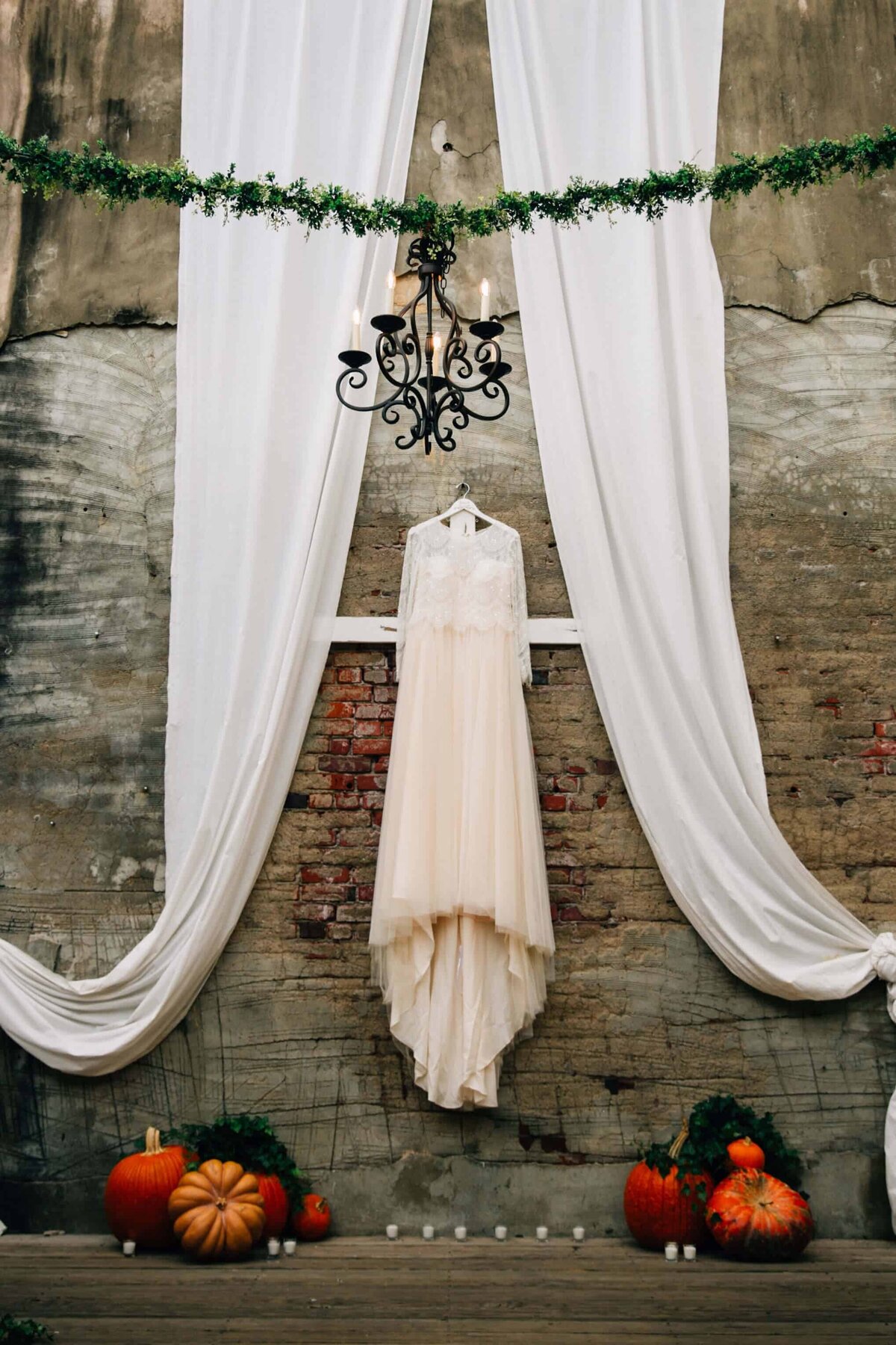 A wedding dress hangs against a brick wall, framed by white drapery. Pumpkins and candles decorate the wooden landing in front of the wall. A black chandelier hangs on green garland above.