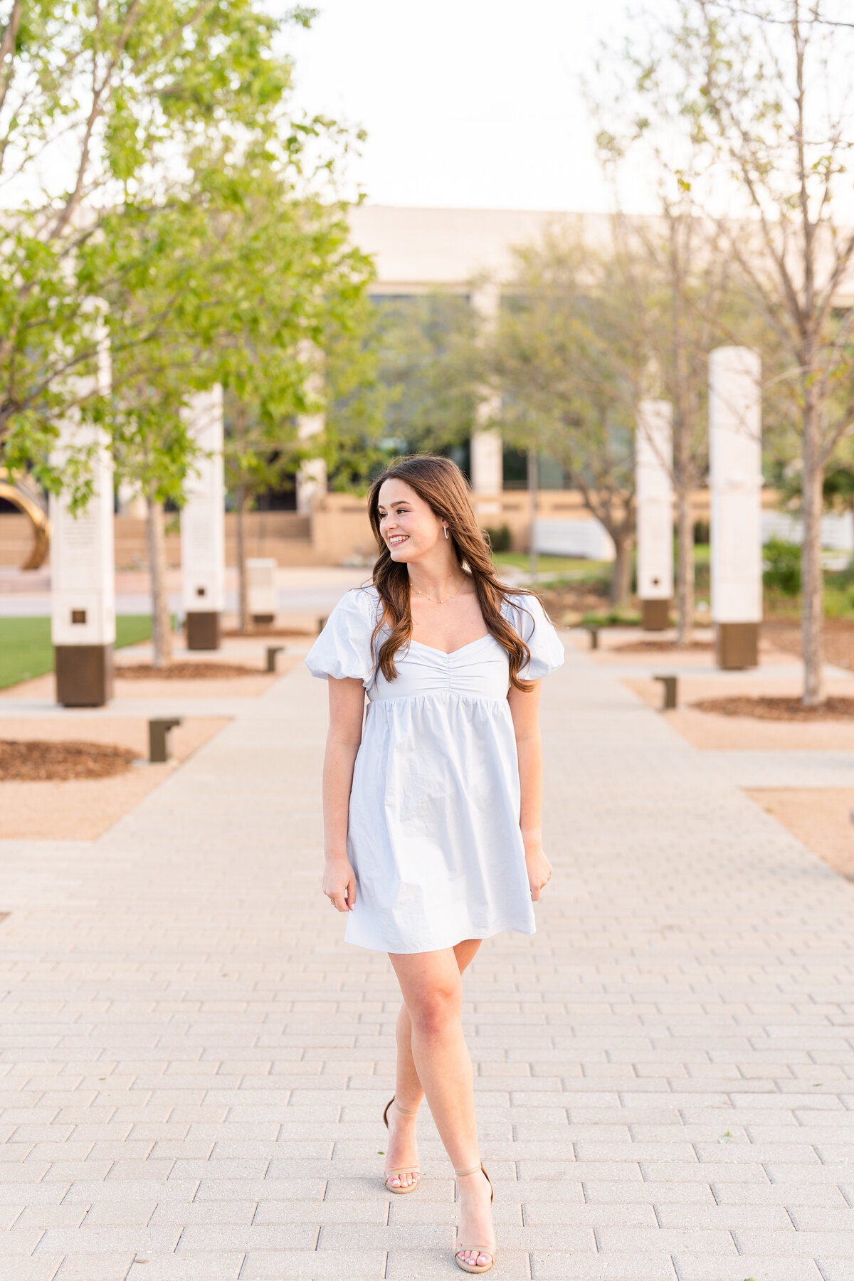 Texas A&M senior girl walking and laughing over shoulder while wearing blue dress in Aggie Park