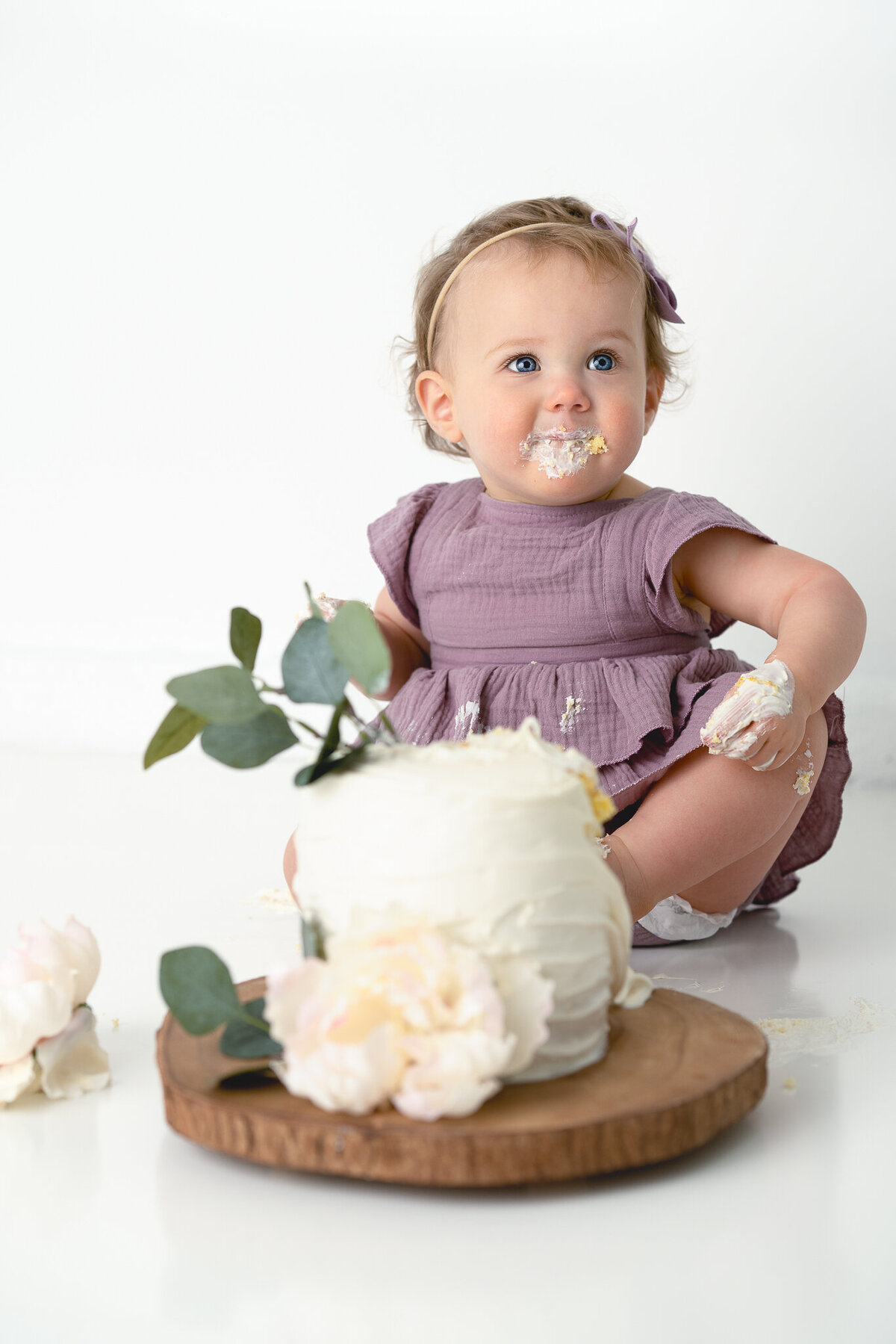 A baby girl is sitting in front of a white cake at her cake smash photography session.