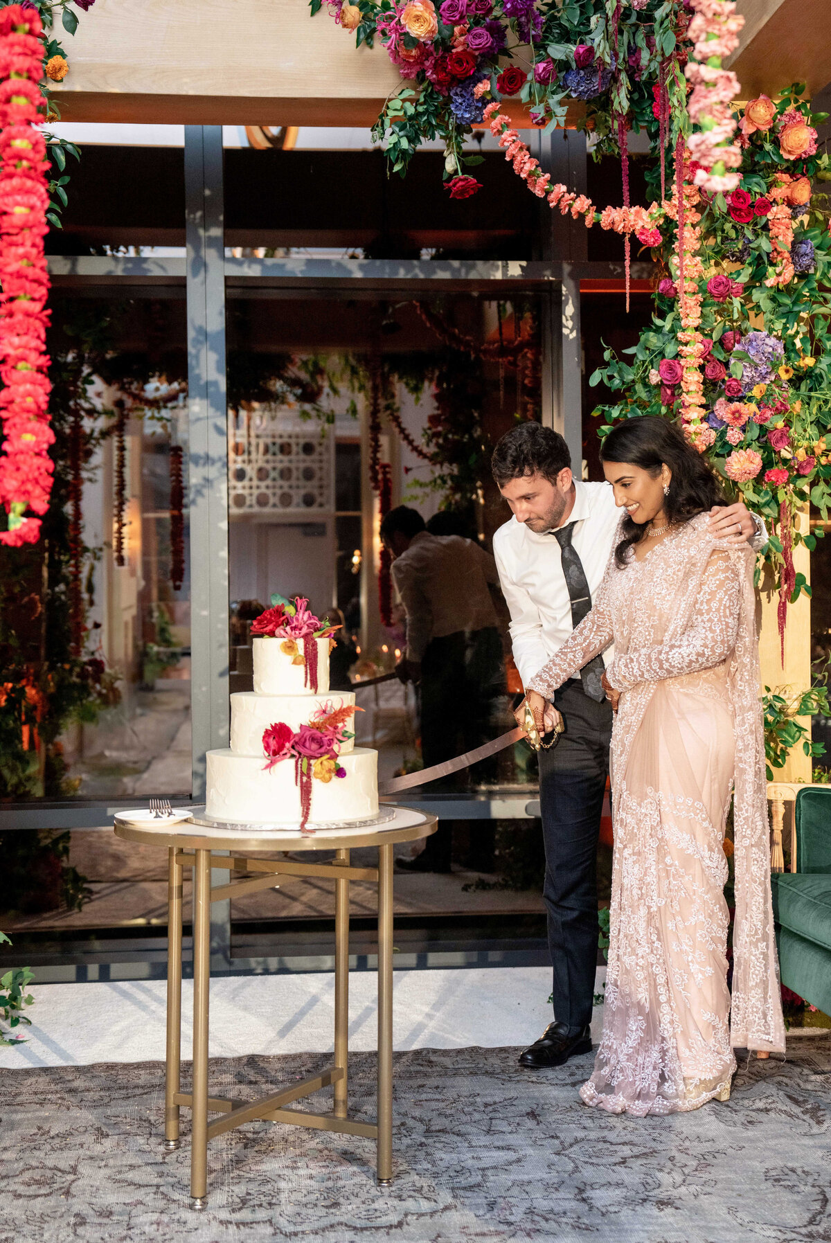 A couple in formal attire cuts a three-tiered wedding cake adorned with flowers. They are surrounded by vibrant floral decorations, and the setting has a festive ambiance with warm lighting.