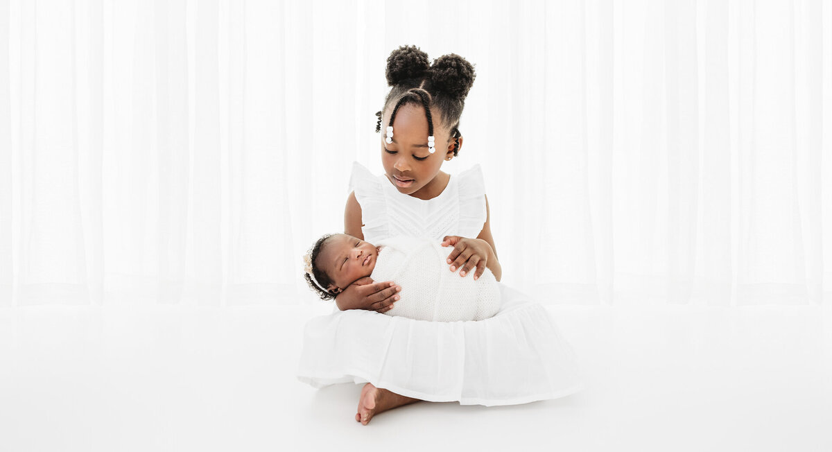 The picture is a serene portrayal of a newborn baby girl being held by her older sister. The setting is a pristine white studio, creating a clean and classical atmosphere. Photographer Bri Sullivan has captured the tender moment with expert precision, highlighting the innocence and bond between the siblings.