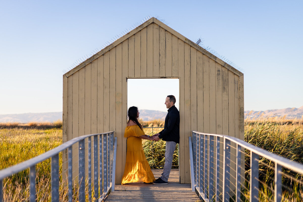 Couple holding hands and smiling at each other inside a wooden structure in a golden marsh – Bay Area Couples Photography Portfolio - Ellobelle Photography