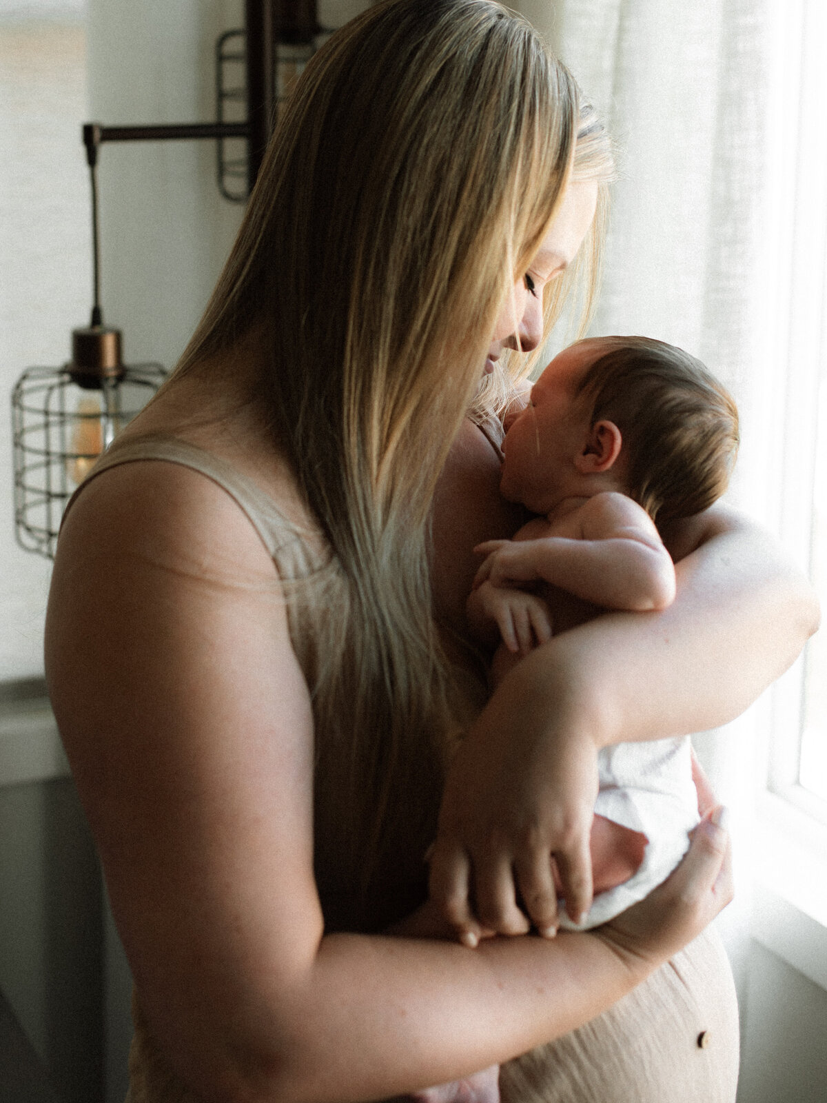 Mother soaking up the morning light while snuggling her newborn near the window inside their home.
