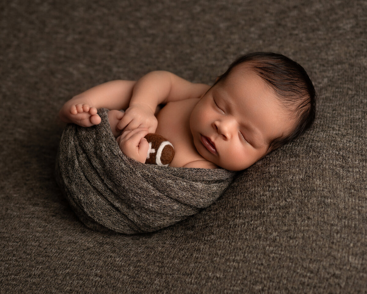 Newborn baby boy in gray wrap holding mini felt football, posed on matching fabric | Studio newborn photography in Austin and Round Rock