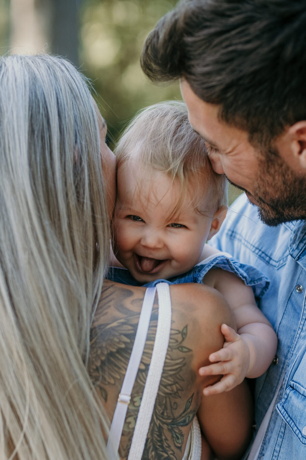 Babygirl puts her tongue out, smiling while being hugged by mom and dad.