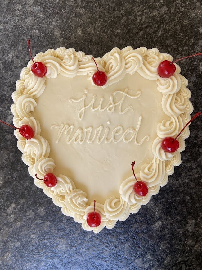 A top down shot of a white heart-shaped cake with fancy piping around the edges, maraschino cherries, and "Just Married" written on top. 