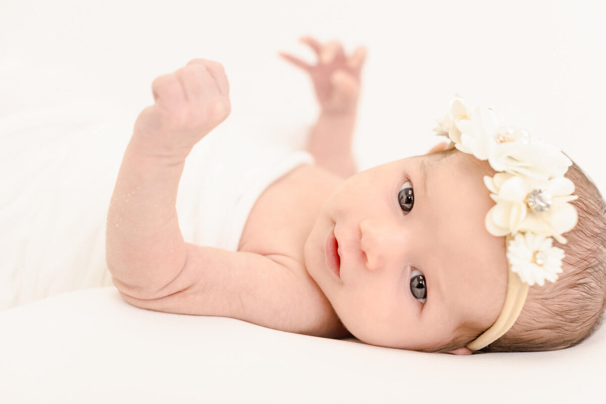 A baby wearing a floral headband lies on a white surface, looking up with wide eyes. The infants left arm is raised, and the background is soft and light, creating a serene atmosphere.
