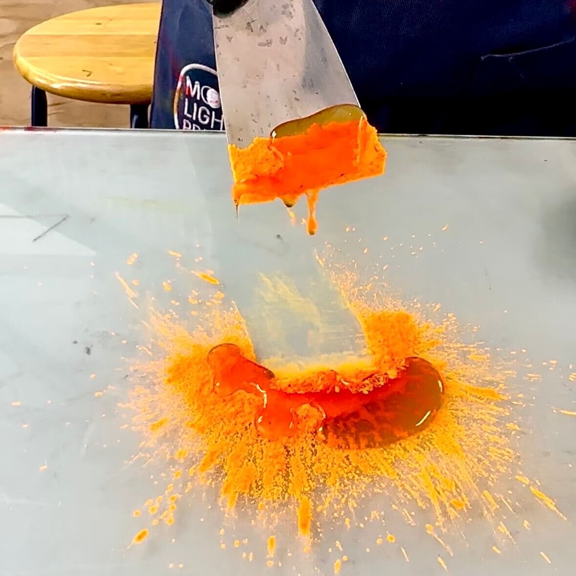 A bold orange pigment bursts out under binder and palette knife, capturing the energetic moment of mixing ink by hand.