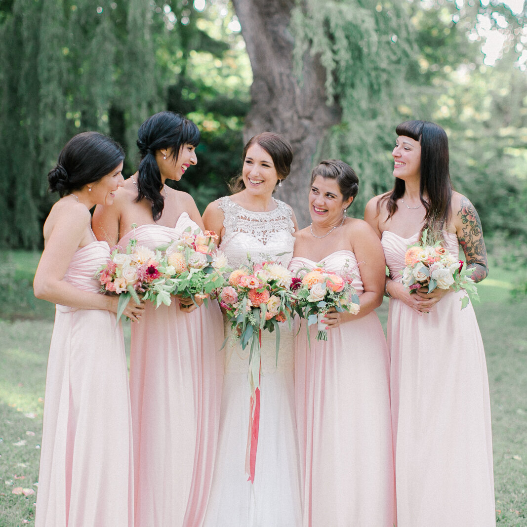 A bridal party stands smiling in a Montreal park by a willow tree, holding lush bouquets.