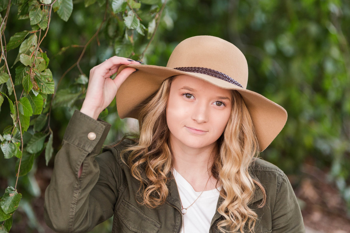 triway high school senior girl wearing olive green jacket over a white tshirt, holding the brim of a big tan hat, photographed in front of gorgeous tree at the OARDC Wooster, photo is only waist up.  Photographed by Jamie Lynette Photography Canton Ohio Senior Photographer