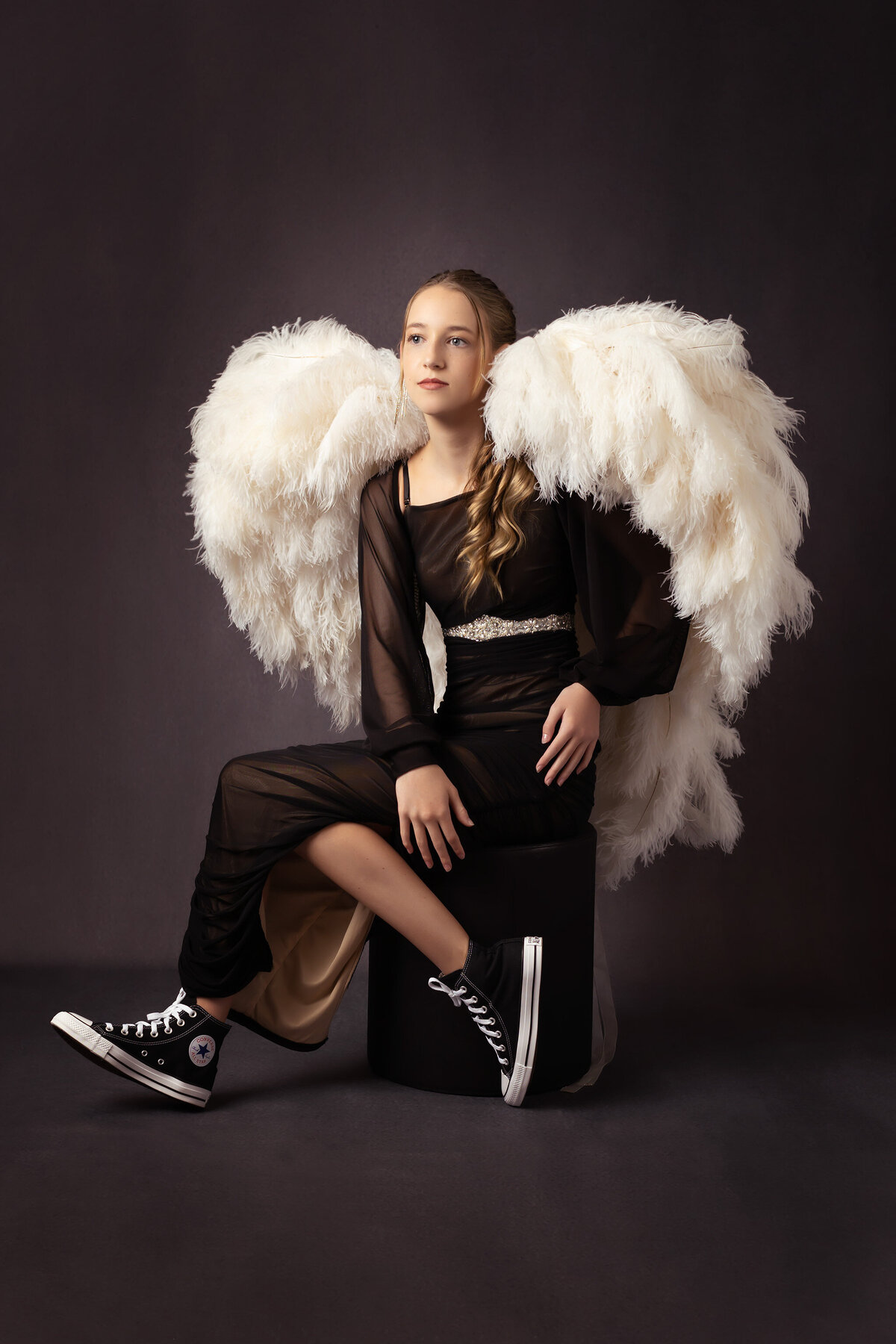 A senior in a sheer black dress with large angel wings sits on a black stool, sporting Converse sneakers, exuding a unique and edgy vibe. Captured by a Dallas senior photographer.