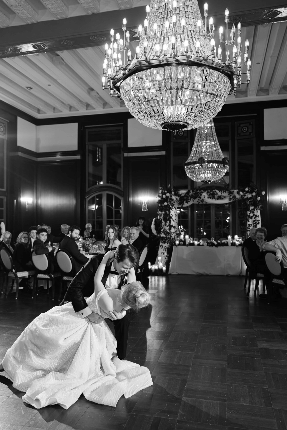 Black and white photo of a bride and groom sharing a romantic dip during their first dance beneath a grand crystal chandelier. Surrounded by seated guests and an elegantly decorated sweetheart table with floral arrangements and candlelight, the moment captures timeless elegance and celebration in a formal ballroom setting.