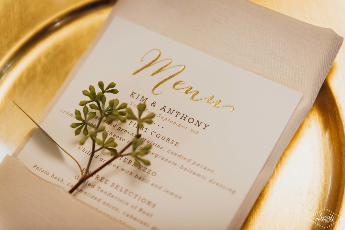 place setting with gold charger plates, ivory napkin, printed menu card, and seeded eucalyptus napkin treatment