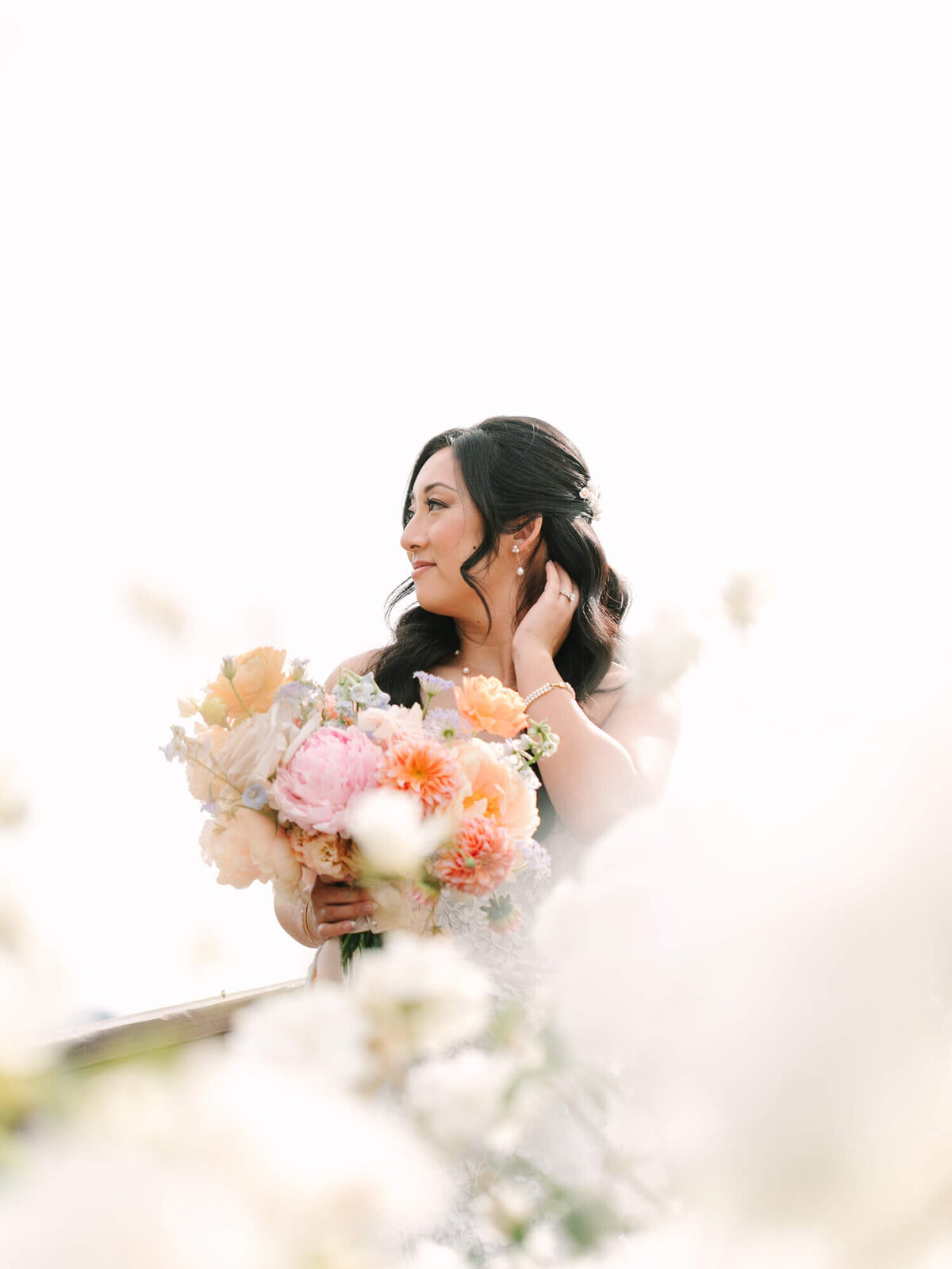 Woman with long dark hair holds a bouquet of pastel flowers, looking serene. Soft focus and bright light give a dreamy, romantic feel.