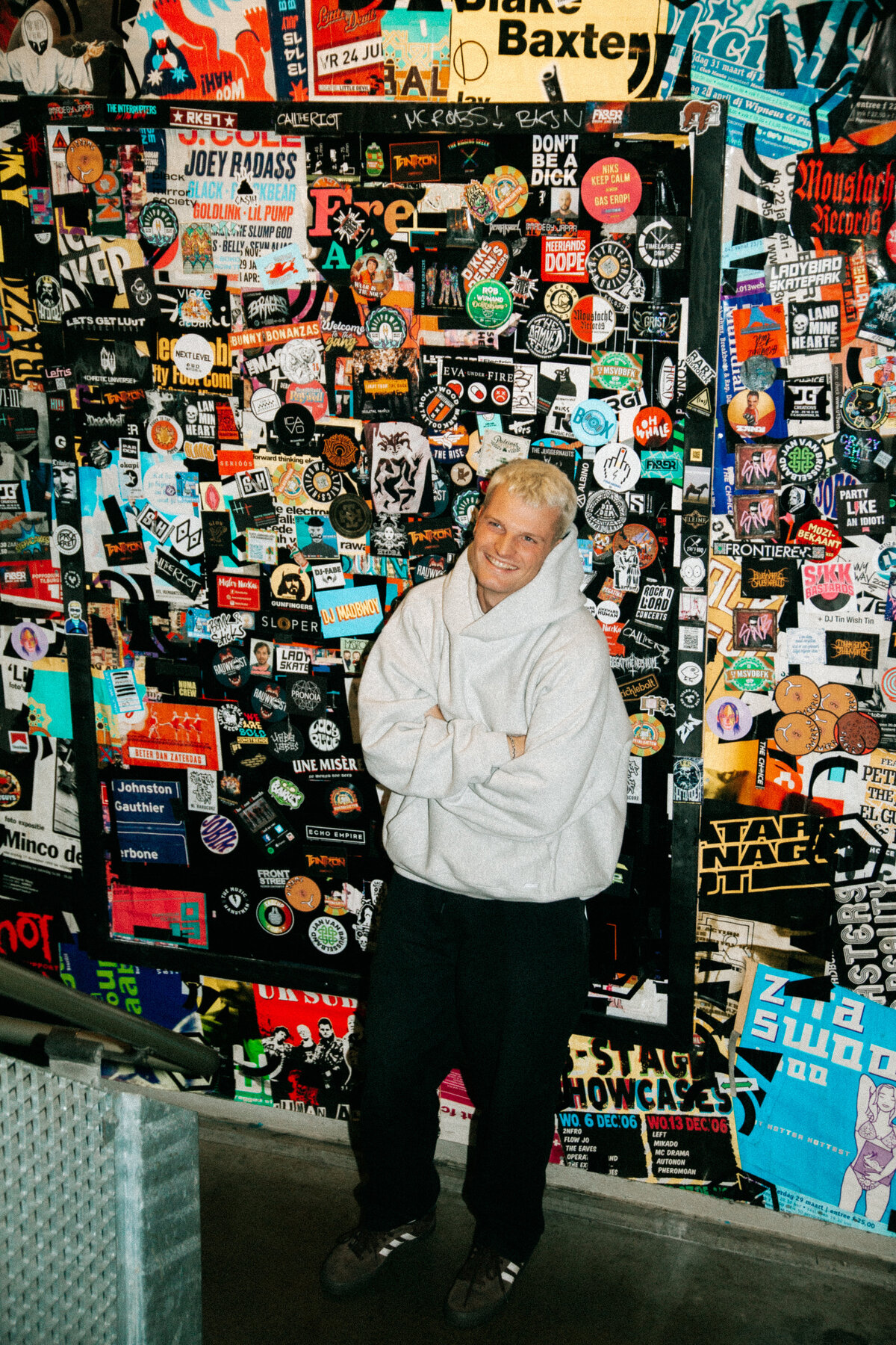 Sam Hofman posing in front of a wall covered with colorful stickers and posters backstage during his club tour, captured by Bo Eppink.